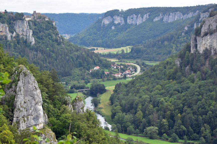 Blick aus großer Höhe auf einen Abschnitt des Oberen Donautals mit bewaldeten Berghängen, die links und rechts der schlingenförmig verlaufenden Donau aufsteigen. Aus den Hängen ragen zudem steile Felszacken heraus.