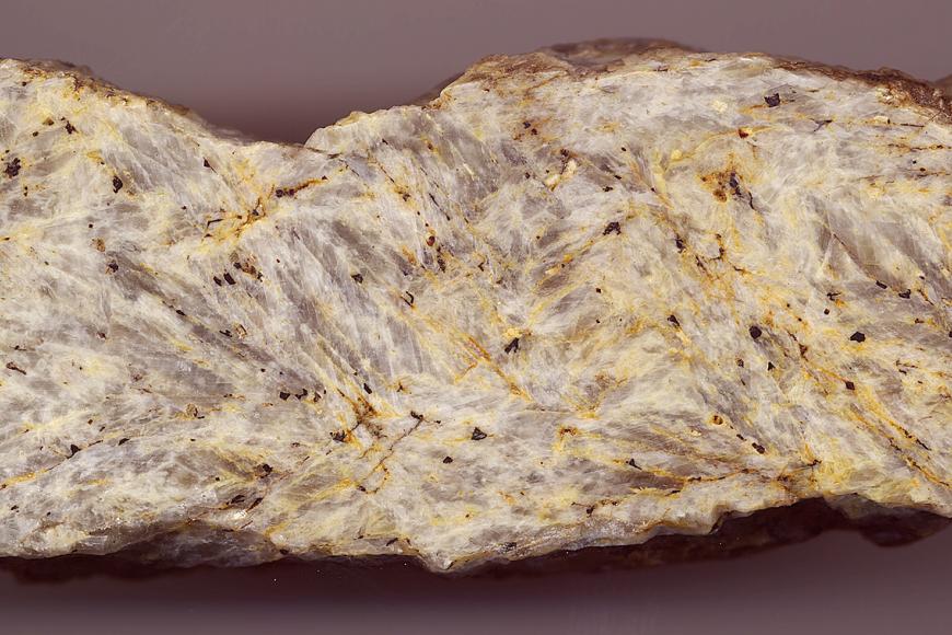 Großaufnahme eines länglichen Steins mit teils abgerundeten Kanten. Die glattgeschliffene Schauseite ist weißlich grau mit gelben Sprenkeln.