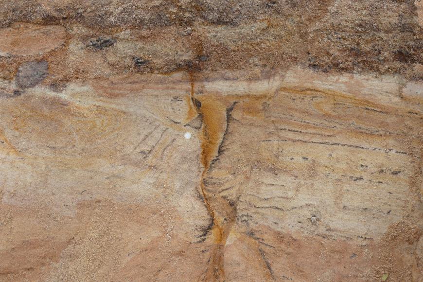 Nahaufnahme von hellbraunem Sand, überkront mit dunkleren, rötlich braunen Schichten. In der Mitte des Bildes verläuft eine senkrechte, orange Linie mit keilförmiger Ausprägung am oberen Ende. 