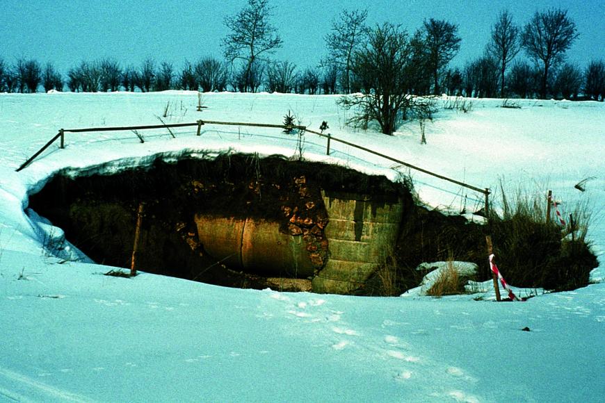 Das Bild zeigt eine schneebedeckte Landschaft. In der Bildmitte ist eine dunkle Senke im Boden, die von einem Zaun umschlossen wird.