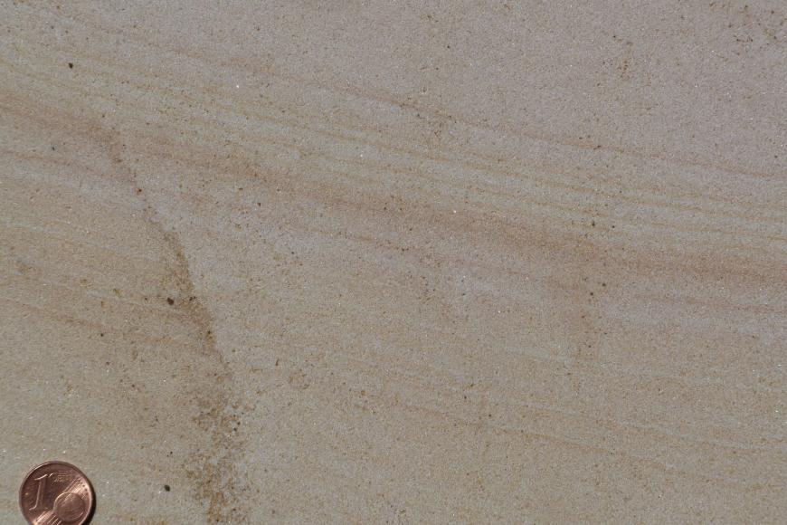 Nahaufname einer Gesteinsplatte mit geschliffener Oberfläche. Farbe gelblich braun, mit nach rechts abfallenden dunkleren Streifen. Eine Cent-Münze links unten dient als Größenvergleich.