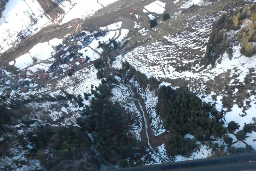 Blick aus dem Fenster eines Hubschraubers auf einen teilweise schneebedeckten Hang mit einigen Bäumen. Der Hang ist von einem Bachbett durchschnitten. Am oberen Ende des Hanges ergießt sich braunes Erdreich in den Bach.