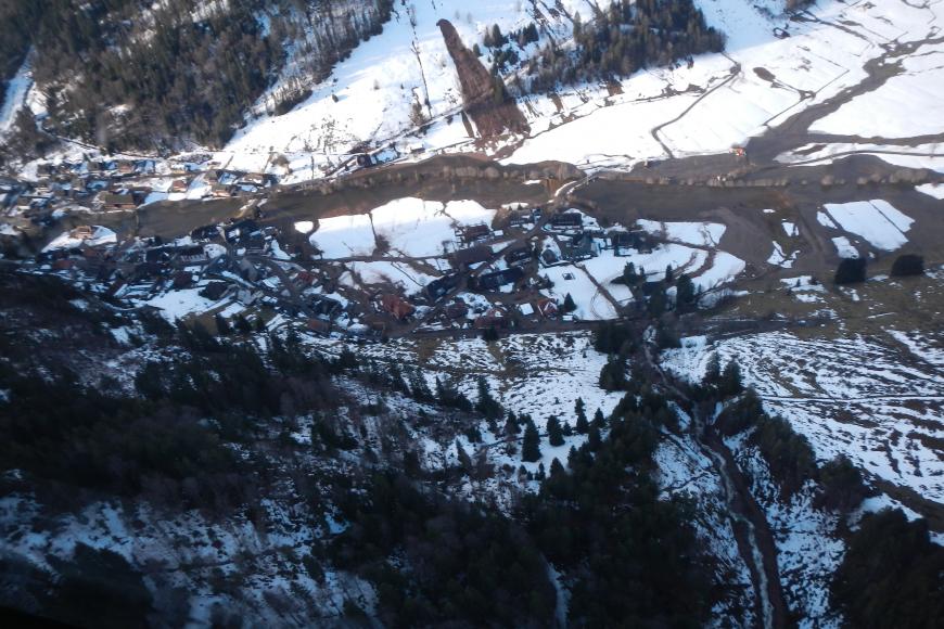 Blick aus der Luft auf ein Tal mit Ortschaft, durch die ein Fluss fließt. An den sich gegenüberliegenden, teilweise schneebedeckten und teils bewaldeten Hängen lassen sich am unteren Bildende ein Murgang, am oberen Bildende eine weitere Hangmure erkennen.