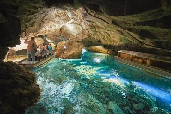 Blick in das Innere einer Höhle. Der Boden ist mit grünlichem Wasser bedeckt, das einen See bildet. Die gewölbte Decke ist braun, wo Licht sie erhellt. Die Höhle hat zwei enge Ausgänge, wovon der linke von einem Stocherkahn mit Fahrgästen benutzt wird.