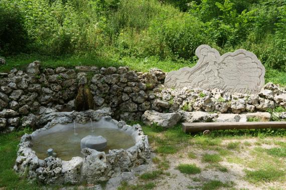 Blick auf eine runde, aus Felsgestein und Beton gefertigte Quellfassung. Der Quelltopf befindet sich links im Bild, vor einer aus Felsblöcken bestehenden Mauer. Rechts davon ist eine Steintafel zu sehen, mit dem Motiv eines Schäfers.