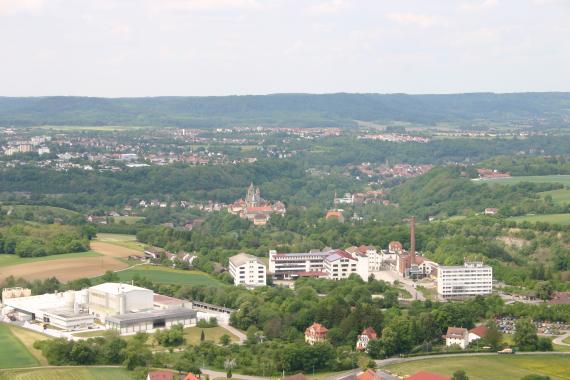 Blick aus großer Höhe über eine weite, hügelige Landschaft, in der sich zwischen ausgedehnten Waldflächen immer wieder Siedlungen zeigen. Im Vordergrund liegen Industrieanlagen. Im Hintergrund steigen bewaldete Berge mit flachen Rücken auf.