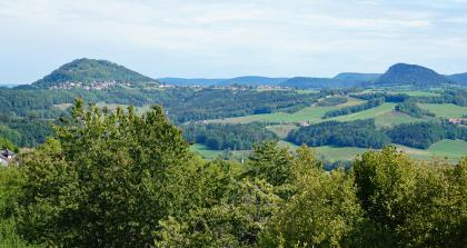 Im Osten des Hohenstaufens erblickt man die beiden anderen „Kaiserberge“ Rechberg und Stuifen. Die Bergkegel aus Oberjuragestein erheben sich beide auf einem schmalen von der Eisensandstein-Formation gebildeten Plateau. Blick von erhöhtem Standort über Baumipfel auf eine hügelige, teils bewaldete Landschaft. Im Hintergrund links erhebt sich ein bewaldeter, von einer Ortschaft umgebener Bergkegel. Rechts außen sind zwei weitere, abgestufte Berge zu erkennen.
