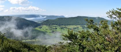 Blick vom Breitenstein nach Südwesten ins Lenninger Tal und nach Westen zur Teck Von erhöhtem Standort blickt man in diesem Panoramabild auf mehrere Bergrücken, die teils bewaldet sind, teils grüne Hänge aufweisen. Links im Hintergrund befindet sich ein Taleinschnitt mit tiefhängenden Wolken. Im Vordergrund wachsen Sträucher.