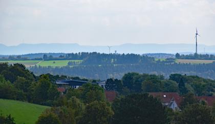 Blick von der Königskanzel über die Heckengäu-Flächen im Oberen Muschelkalk nach Südosten zur Westlichen Schwäbischen Alb Blick aus größerer Höhe über eine bewaldete Hügellandschaft. Im Vordergrund, zwischen Bäumen, liegen die Häuser einer Ortschaft. Dahinter folgt eine Senke mit bewaldetem Gegenhang. Im Hintergrund verlaufen flachstufige bewaldete Berge.