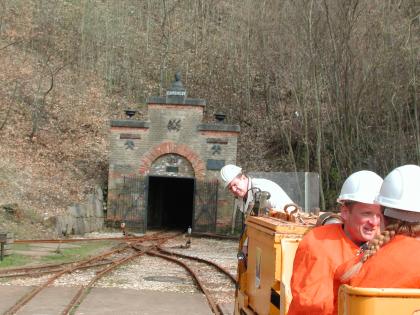 Besucherbergwerk Tiefer Stollen in Aalen-Wasseralfingen Blick auf den geöffneten, an einem Waldhang gelegenen Eingang eines Bergwerks. Im Vordergrund liegen Schienen der Grubenbahn.
