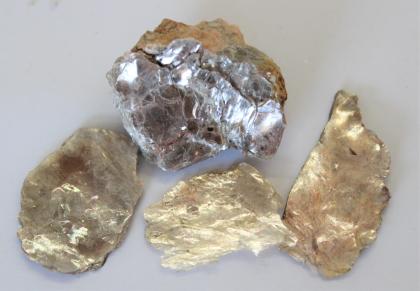 Muskovit aus Pegmatitgang im Wildbader Granit Großaufnahme von vier Gesteinsstücken. Die untere Reihe zeigt drei Handstücke in Gold- und Bronzetönen, darüber liegt ein etwas größeres, teils silberfarbenes Stück.