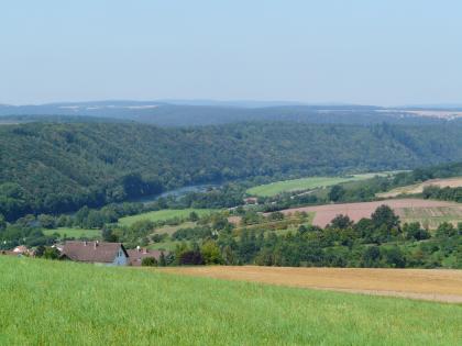 Blick über hochgelegene Wiesen und Felder auf ein Flusstal. Auf der linken Seite des Flusses, zum Hintergrund hin, erheben sich bewaldete Bergrücken. Rechts unterbrechen Ackerflächen und Wiesen die Bewaldung.