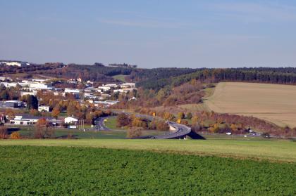 Westrand des Baulands bei Buchen Blick von hochgelegenen, begrünten Äckern auf Industriebauten links mit Straßenanschluss sowie weiteren Ackerflächen rechts. Waldgebiete trennen beide Bereiche und setzen sich auch im Hintergrund fort.