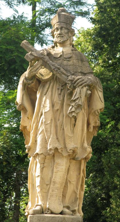 Brückenheiliger auf der Nepomukbrücke in Bad Krozingen Das Foto zeigt eine größere, aus gelblich grauem Gestein gefertigte Heiligenfigur. Die Figur hält ein Kreuz mit Christus in den Händen. Im Hintergrund stehen hohe Bäume.