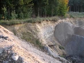 Blick über einen nach rechts geneigten, offenen Hang. Auf der rechten Seite des gelblich braunen Hanges ist grauer Sand aufgehäuft. Die obere Kante des Hanges ist bewaldet.
