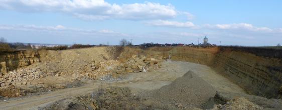 Panoramabild von ringförmig verlaufenden, niedrigen Steinbruchwänden. Im Vordergrund sowie auf der linken Bildhälfte sind Schutthalden zu sehen. Die rechte Steinbruchwand liegt im Schatten.