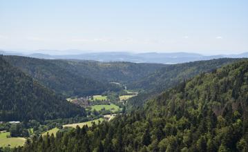 Blick aus großer Höhe in ein kleines, teilweise besiedeltes Tal. Zu beiden Seiten sowie am Ende des Tales steigen bewaldete Berghänge auf. In der Ferne sind weitere, höhere Berge zu erkennen.
