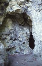 Blick auf ein aus graublauem Gestein gebildetes Felsentor, das den Eingang zu einer sich rechts nach einer Felswand öffnenden Höhle freigibt.