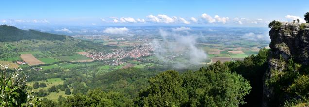 Aus großer Höhe blickt man in diesem Panoramabild auf eine weite Landschaft mit Feldern, Wäldern und Siedlungen. Rechts erhebt sich eine markante, vorspringende Felsengruppe als Aussichtspunkt. Links im Mittelgrund befindet sich ein bewaldeter Bergrücken.