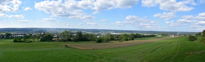 Weiter Blick über flachhügelige, nach rechts hin leicht ansteigende Wiesen und Äcker. Im Hintergrund Bauminseln, Ortschaften und langgezogene bewaldete Bergrücken.