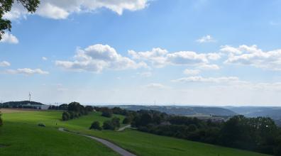 Blick von einem Aussichtspunkt auf einen nach rechts abfallenden grünen Hügel mit Baumbestand. Rechts im Hintergrund folgt eine stufige Landschaft mit flachen Rücken und Senken sowie bewaldeten Bergen.