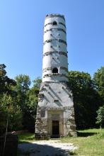 Blick auf einen gemauerten Aussichtsturm. Der Turm ähnelt einem verbreiterten Schornstein mit weiß gestrichenen Wänden und grauen Mauerkränzen. Der Turm steht auf einer Waldlichtung.