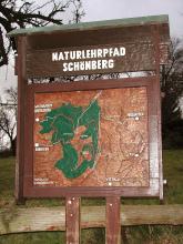 Blick auf eine hölzerne, überdachte Schautafel, die auf den Naturlehrpfad am Schönberg hinweist. 