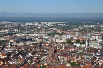 Von sehr hohem Standort blickt man über die ausgedehnten Häuserzeilen einer größeren Stadt. In der unteren Bildmitte erhebt sich ein gotisches Münster. Im dunstigen Hintergrund ist eine Bergkette erkennbar.