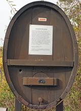 Geologischer Weinlehrpfad Achkarren – Station am Beginn des Lehrpfads Frontansicht eines alten ovalen Weinfasses aus rotbraunem Holz. Im oberen Teil ist eine kleine Hinweistafel sowie ein Richtungspfeil angebracht.