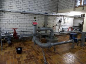 Blick in das Innere eines Gebäudes mit gekachelten Wänden und Fliesenboden. Verschiedene, miteinander verbundene Rohrleitungen sind hier installiert.