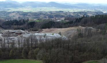Aus großer Höhe blickt man auf eine in ein Waldgebiet eingeschnittene, ausgedehnte Baustelle. Das aufgegrabene Erdreich weist graue, aber auch rötlich braune Farbtöne auf. Zum Hintergrund hin folgen Äcker und Wiesen sowie bewaldete Bergketten.