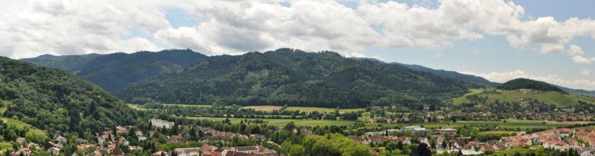 Panoramabild einer besiedelten, grünen Landschaft mit hohen bewaldeten Bergrücken links vorn sowie im Hintergrund. Rechts erhebt sich ein niedrigerer, nur auf der Kuppe bewaldeter Berg. 