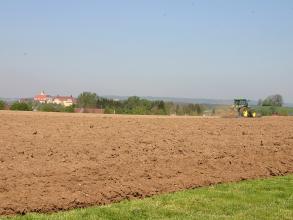 Blick auf einen rötlich braunen, mit Steinen durchsetzten Acker, der von einem Traktor befahren wird. Im Hintergrund Bäume und Gebäude.