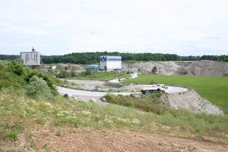 Blick auf eine Kiesgrube mit Gebäuden, Förderbändern, einer kurvigen Straße und Fahrzeugen. 