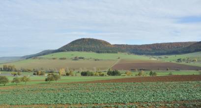 Eine bewaldete und teils langgestreckte, teils runde Erhebung bildet den Mittelpunkt des Fotos. Darunter dehnen sich Hänge mit Grünland- und Ackerflächen aus, durchquert von Straßen. Im Vordergrund ein bepflanzter Acker.