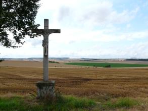 Von einem Wegkreuz links blickt man auf einen braunen, abgeernteten flachen Acker, der bis zum Mittelgrund reicht. Dahinter schließen sich weitere, nun leicht wellige Ackerflächen an.