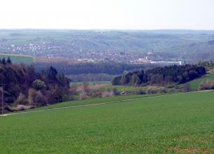 Blick über eine nach links abfallende Grünfläche mit Waldflächen im Mittelgrund. Im Hintergrund, nach einem wieder aufsteigenden Hang, liegen eine größere Ortschaft sowie teils bewaldete Höhen.