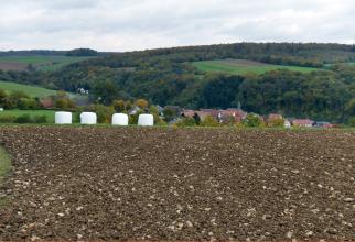 Blick über einen sehr steinigen braunen Acker auf eine von bewaldeten Hügeln und Hochflächen eingeschlossene Ortschaft.
