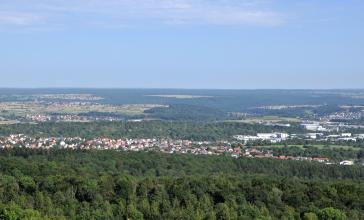 Von erhöhtem Standort blickt man auf eine weite Ebene mit zahlreichen Siedlungen und ausgedehnten Waldflächen. Links und im Hintergrund sind auch Inseln mit Äckern erkennbar.