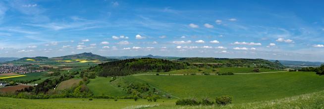 Panoramaansicht von erhöhtem Standort über die Landschaft des Hegaus mit mehreren Bergkegeln. Neben Wiesen und bewaldeten Kuppen finden sich auch Äcker und Siedlungen sowie mehrere Wanderwege.