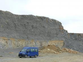 Blick auf eine hohe Abbauwand in einem Steinbruch. Das untere Viertel besteht aus gelblich-beigen gebankten Gesteinen, darüber befindet sich dunkelgraues bis blaugraues Gestein. Vor der Wand steht ein blauer Van.