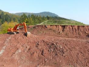 Im Vordergrund des Bildes sieht man eine Abbauwand einer Tongrube. Das rötliche Material wird gerade mit einem Bagger am linken Bildrand abgebaut. Im Hintergrund befinden sich bewaldete Hügel.
