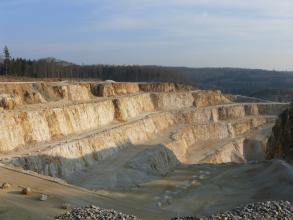 Blick von seitlich oben in einen Steinbruch, in welchem gelbliches Gestein auf mehren Stufen abgebaut wird. Das Gestein leuchtet in der tiefstehenden Sonne, die vordere Hälfte des Steinbruchs liegt im Schatten.