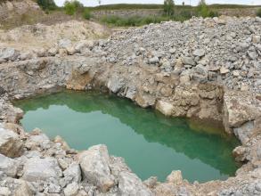 Blick von schräg oben in einen Steinbruch, in welchem hellgrau-beiges Gestein abgebaut wird. In der Bildmitte und der untersten Sohle des Steinbruchs befindet sich grünliches Wasser.