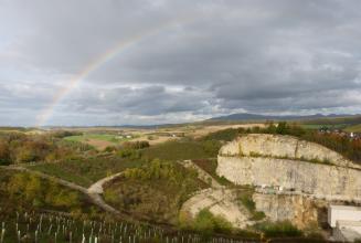 Blick von erhöhtem Standpunkt in einen Steinbruch, welcher sich zur rechten Bildseite hin öffnet. Um den Steinbruch herum befindet sich idyllische Landschaft. Über dem Steinbruch erstreckt sich ein Regenbogen.