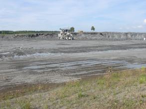 Blick auf eine flache, von Wasser durchzogene graue Bodenfläche. Im Hintergrund erhebt sich eine niedrige Steinbruchwand. Davor ist eine auf Transportschienen laufende Maschine zu sehen. Im Vordergrund folgt von Gras durchsetzter Boden.