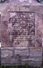 Blick auf den unteren Teil eines steinernen Denkmals mit eingravierten Namen. Das Gestein ist violettgrau und unten längs gestreift.