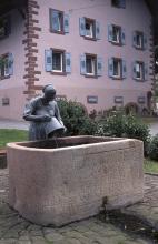 Das Bild zeigt einen rechteckigen, aus rötlich grauem Stein gefertigten Brunnen. In den Stein sind Zeichen und Buchstaben eingeritzt. Eine Frauenfigur mit Wassereimer ergänzt den Brunnen. Im Hintergrund steht ein Gebäude, ebenfalls aus rötlichem Stein.