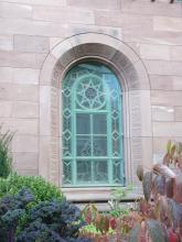 Blick auf ein Rundbogenfenster mit blaugrünen Sprossen und Verzierungen. Das Fenster ist von rötlich grauen Mauersteinen umgeben.