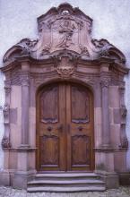 Das Foto zeigt das Eingangsportal einer Kirche mit hölzernen Türen und umlaufenden Verzierungen aus rötlich grauem Stein.